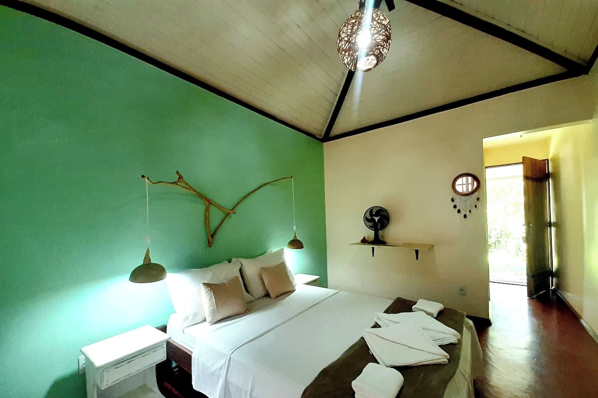 A bedroom with a bed, bedside tables, a hanging light fixture, a wall-mounted shelf, and a window.