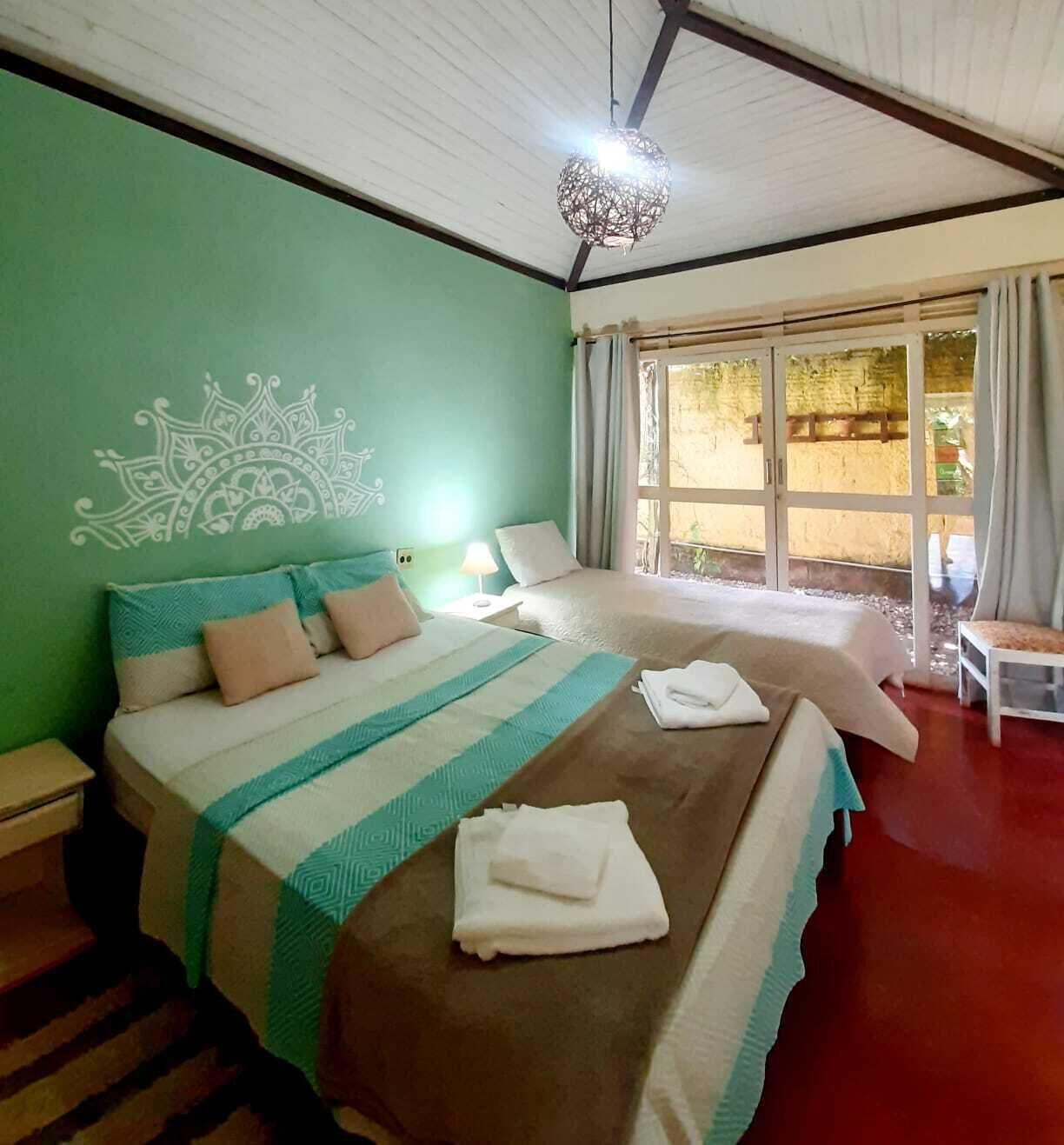 A room with two beds, a decorative wall design, a hanging light fixture, and a view through a glass door.