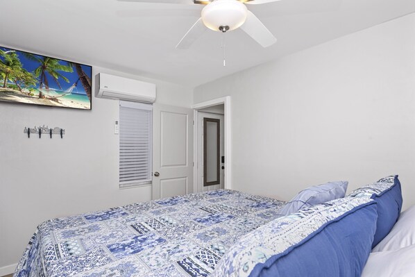 Un dormitorio con una cama, ventilador de techo, unidad de aire acondicionado, una puerta y una obra de arte colgada en la pared.