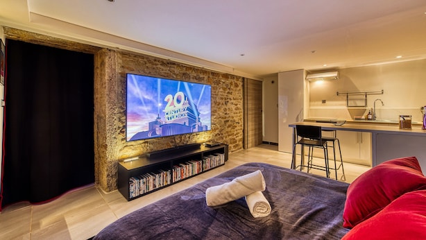 Een moderne woonkamer met een flatscreen televisie op een stenen muur, een klein keukenhoekje, en een knus zithoekje met een plaid en kussens.
