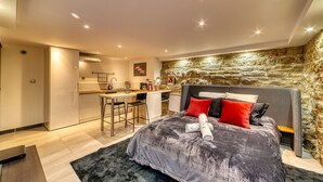 Een moderne slaapkamer met een groot bed, een accentmuur van steen en een kitchenette op de achtergrond.