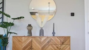 Um armário de madeira com padrão herringbone, com um espelho redondo acima, ladeado por duas plantas em vasos.