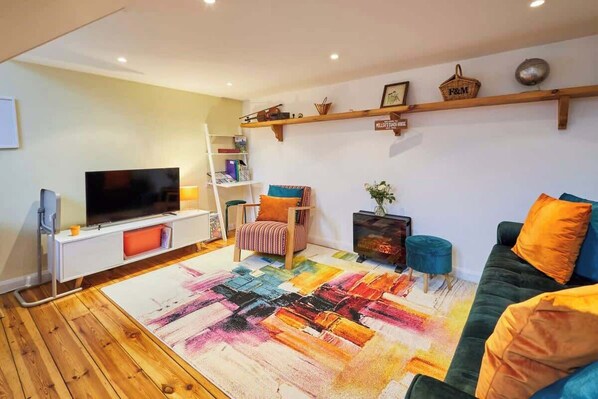 A modern living room with a flat-screen TV, a wooden floor, a colorful rug, a green sofa, and a bookshelf. - Host & Stay | Millers Coach House (Hexham)