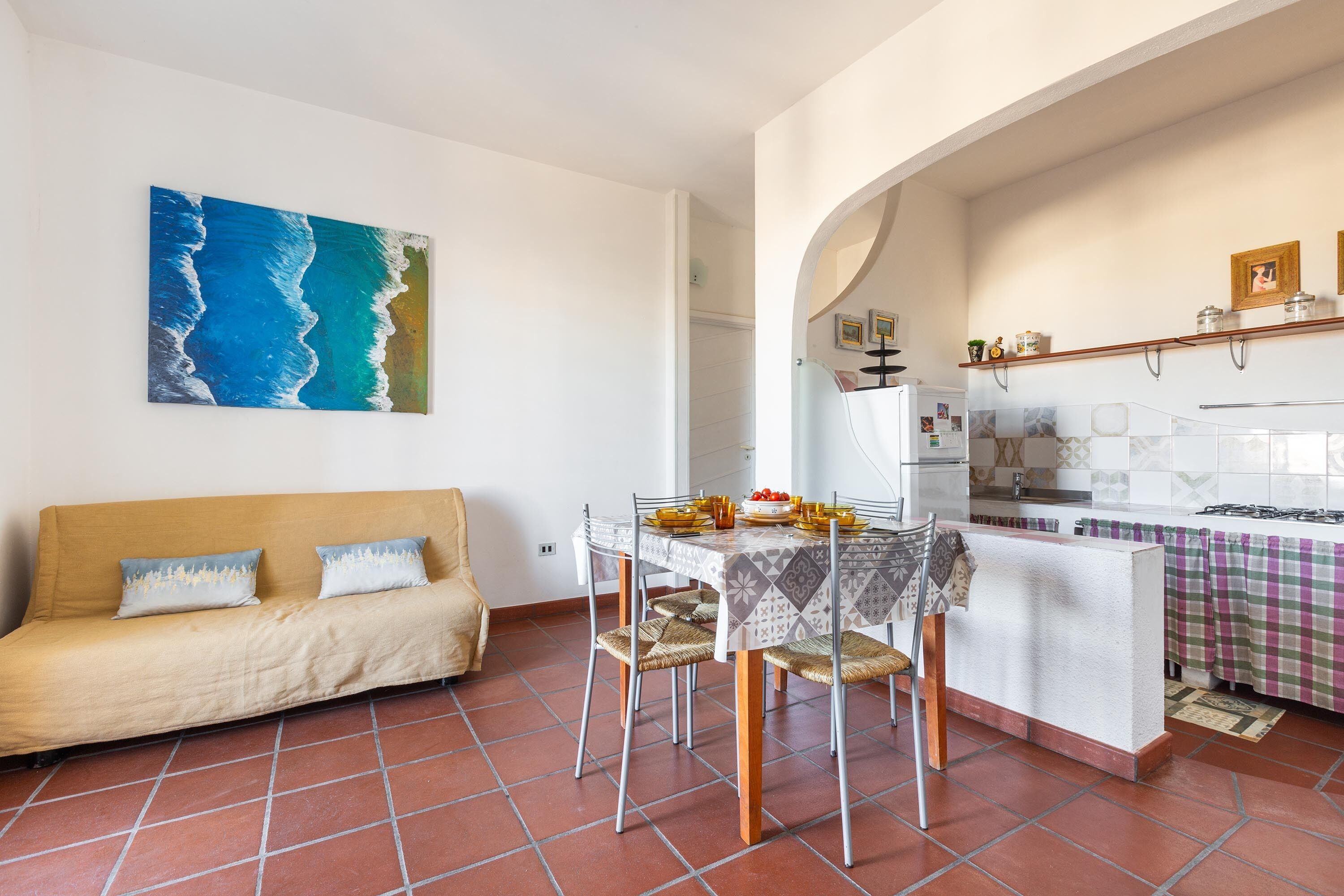 A living room with a sofa, dining table, and chairs, a kitchen with a stove, and a large painting of a beach scene.