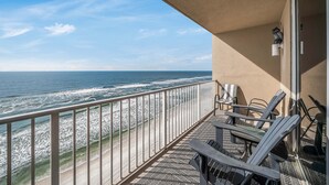 A balcony with a view of the ocean, featuring a metal railing, two Adirondack chairs, and a small table.