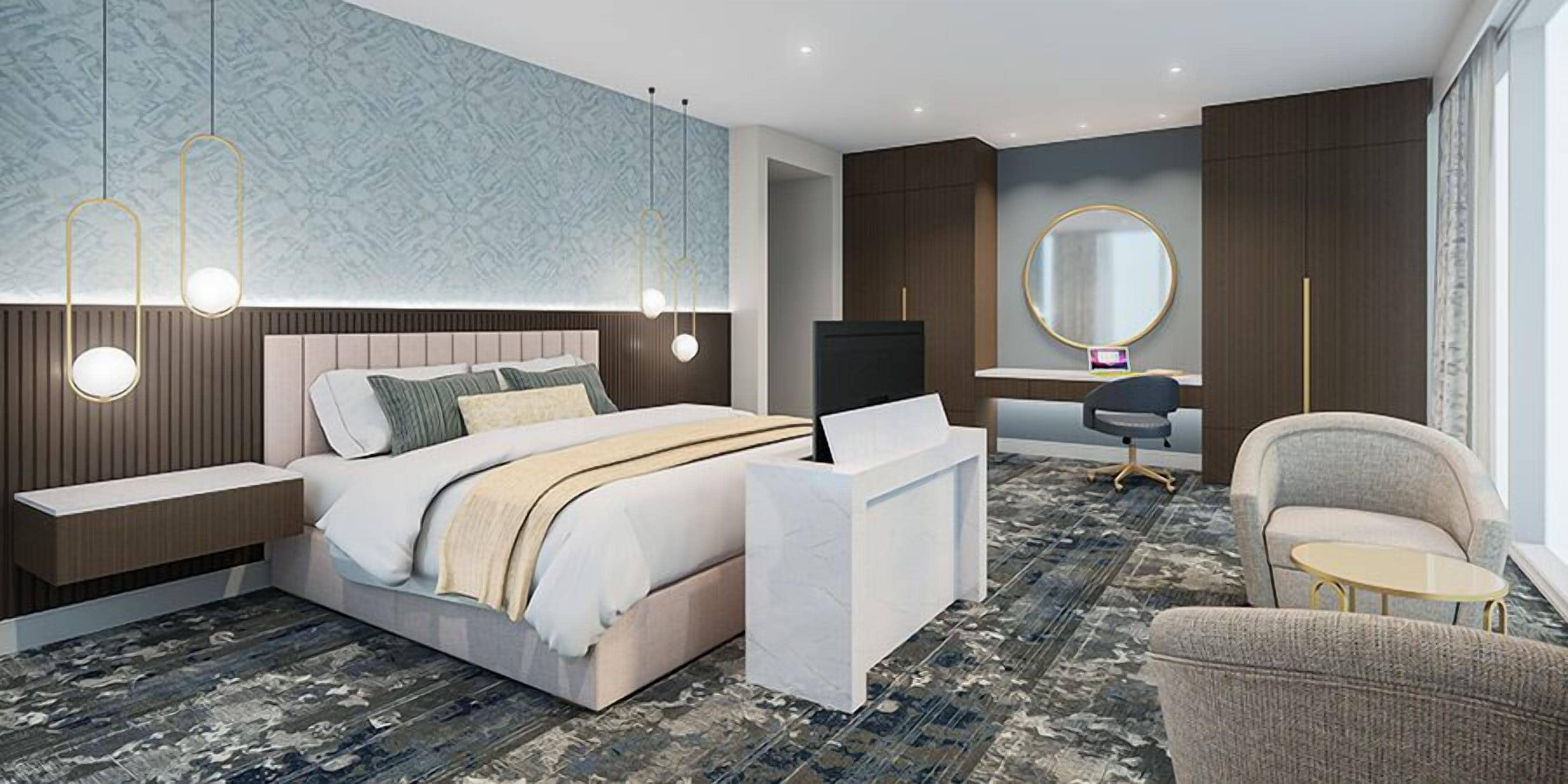 A modern hotel room with a large bed, bedside tables, a desk, a chair, and a round mirror.