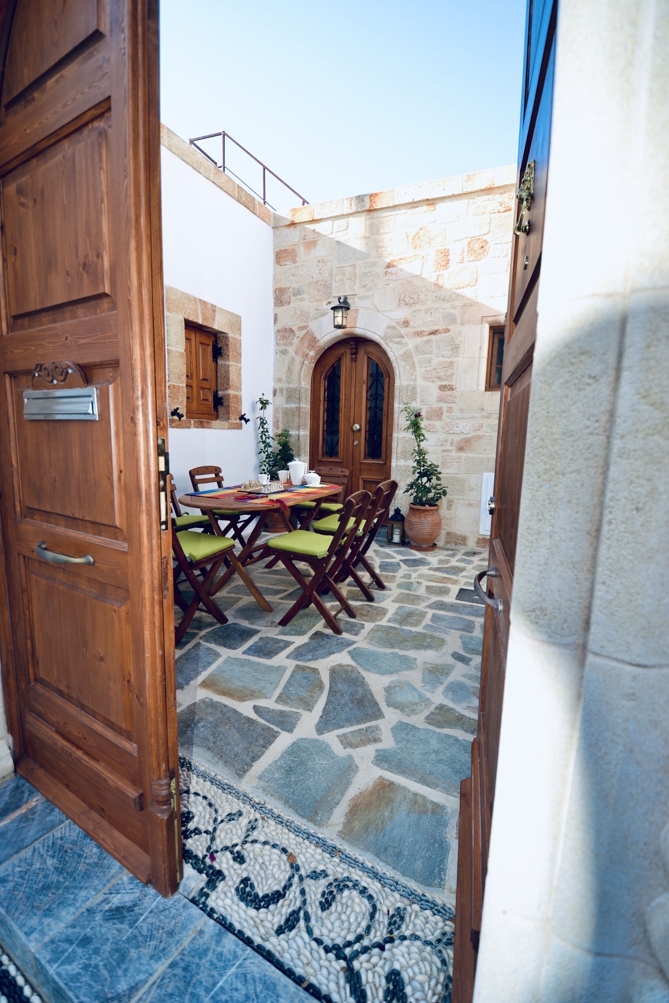 Une porte en bois ouverte donne sur une cour dallée de pierres, où se trouvent une table et des chaises, un pot de fleurs et un mur en pierre.