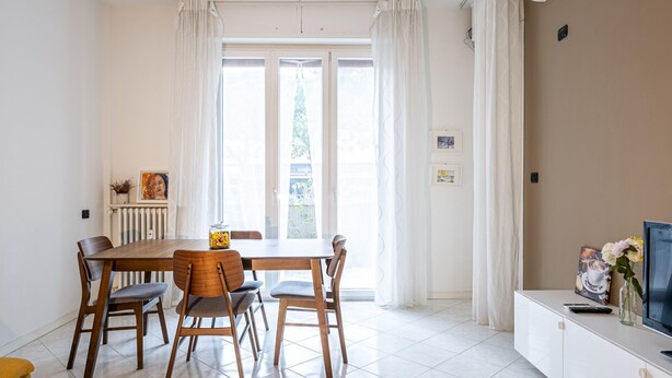 A dining area with a wooden table and chairs, a small vase with flowers, and a TV on a white cabinet.