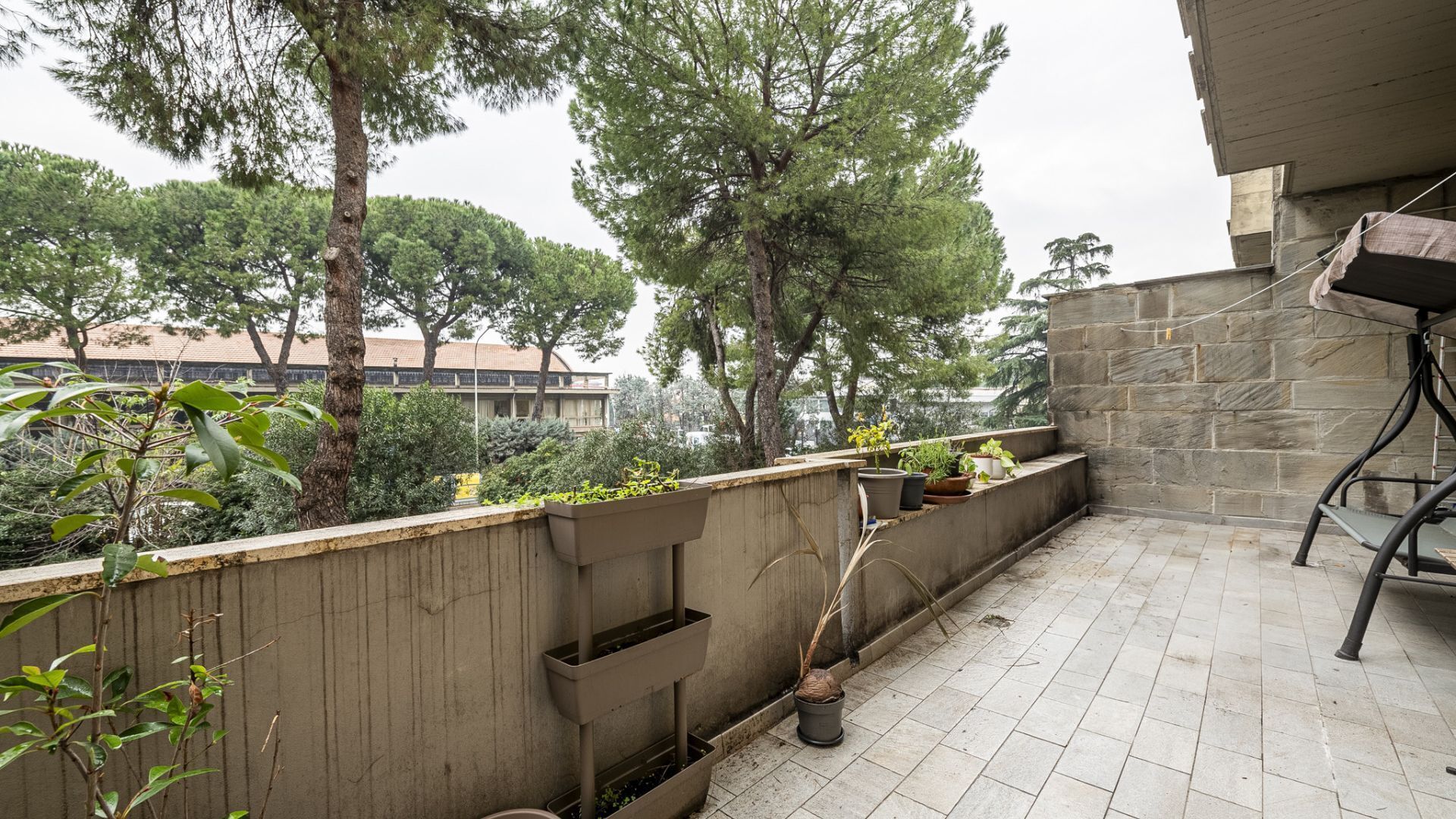 A rooftop terrace with a stone wall, potted plants, and a view of surrounding trees and buildings.