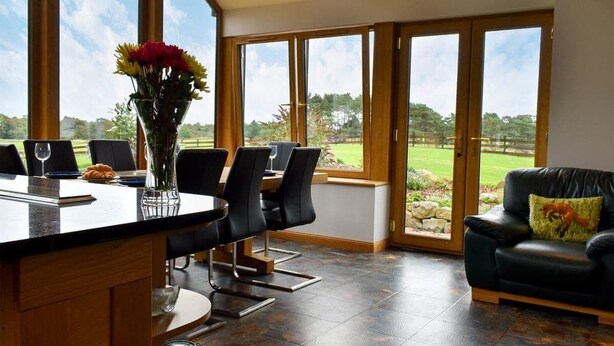 A spacious dining area with a large window overlooking a garden, a black leather sofa, and a glass vase with flowers on the counter. - 2 Bedroom bungalow Aberdeen within the beautiful countryside. (Aberdeen)