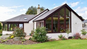 A single-story house with a sloped roof, large windows, and a well-maintained garden with various plants and a lawn. - 2 Bedroom bungalow Aberdeen within the beautiful countryside. (Aberdeen)