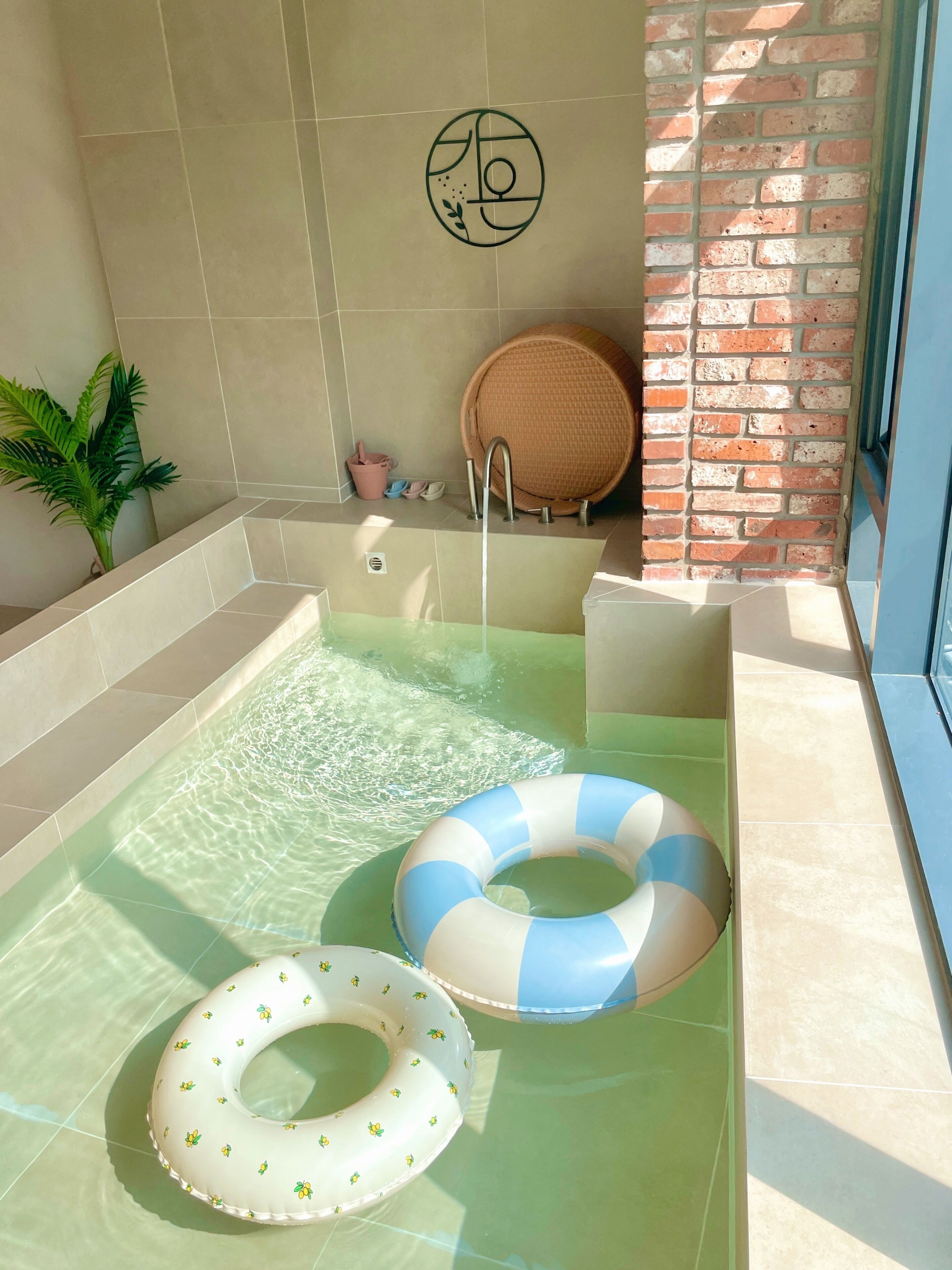 Un área de spa con un jacuzzi, dos anillos hinchables, una planta en maceta y una pared de ladrillo.
