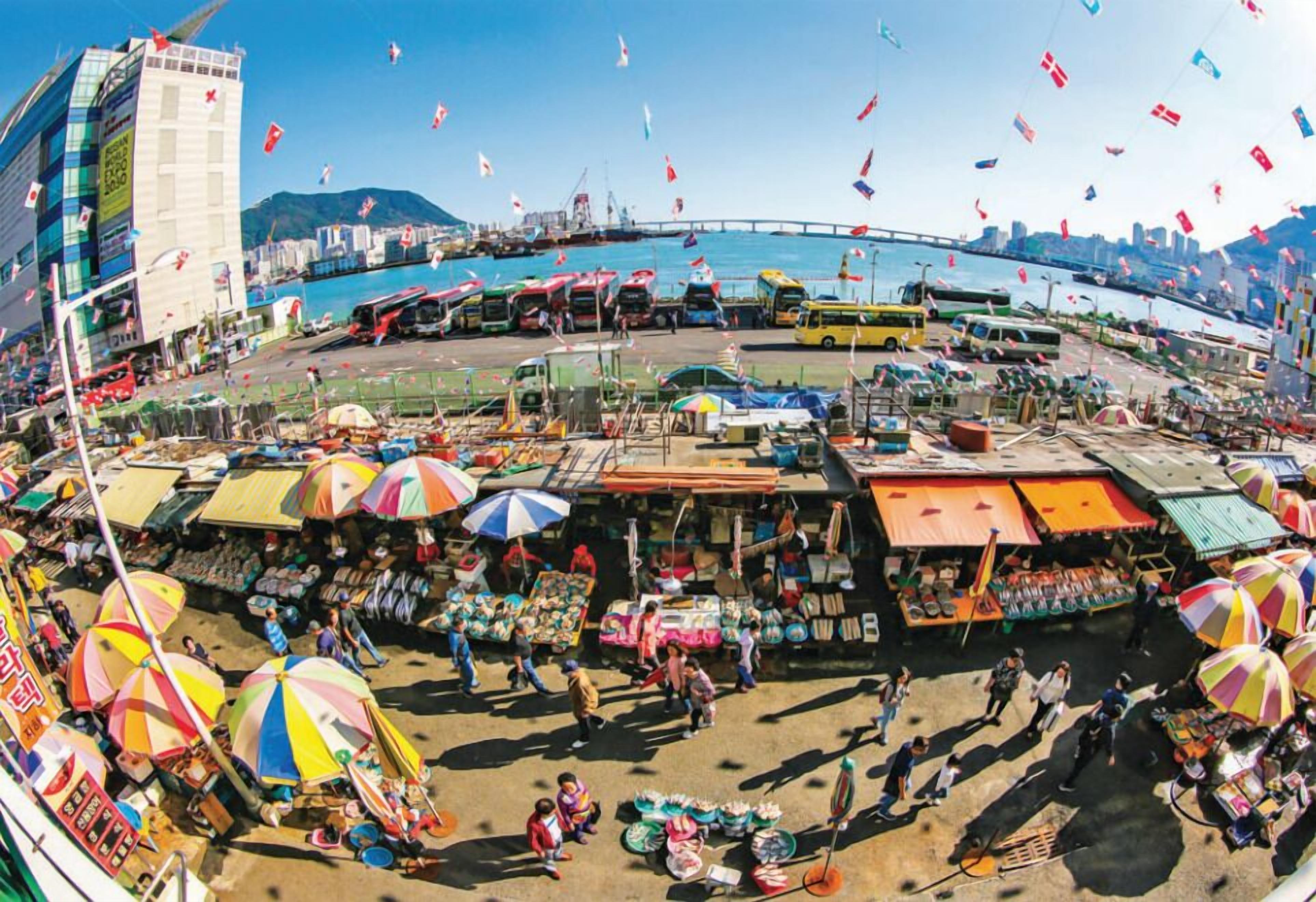 A bustling market area with various stalls and umbrellas, a bridge in the background, and a body of water.
