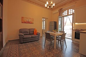 A spacious living area with a sofa, dining table, and chairs. There is a chandelier, a painting on the wall, and a view through a glass door.