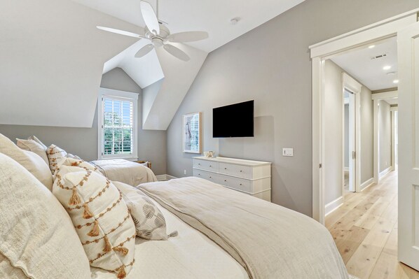 A bedroom with a large bed, a ceiling fan, a TV mounted on the wall, and a window with blinds.