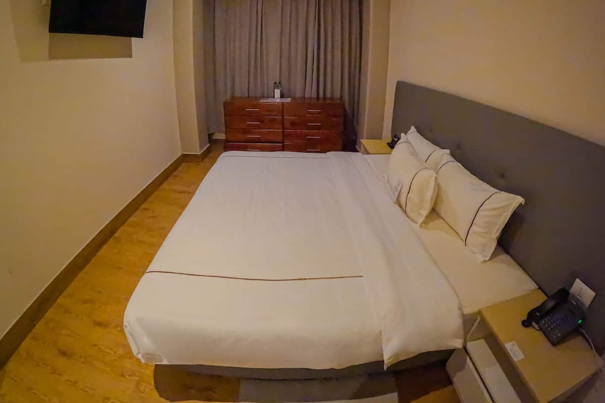 Une chambre d’hôtel avec un grand lit, une commode en bois, un téléviseur à écran plat fixé au mur et une table de chevet avec un téléphone et une boîte.
