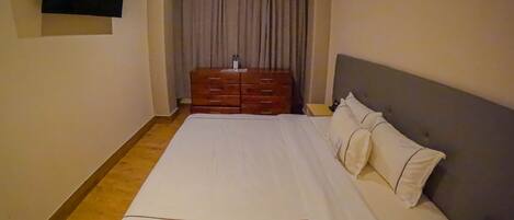 A hotel room with a large bed, a wooden dresser, a flat-screen TV mounted on the wall, and a bedside table with a phone and a box.