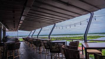 An outdoor dining area with tables and chairs, overlooking a body of water and greenery.