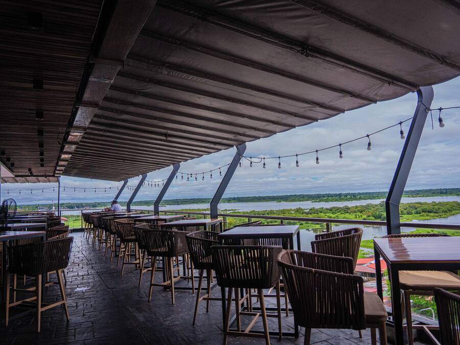 An outdoor dining area with tables and chairs, overlooking a body of water and greenery.
