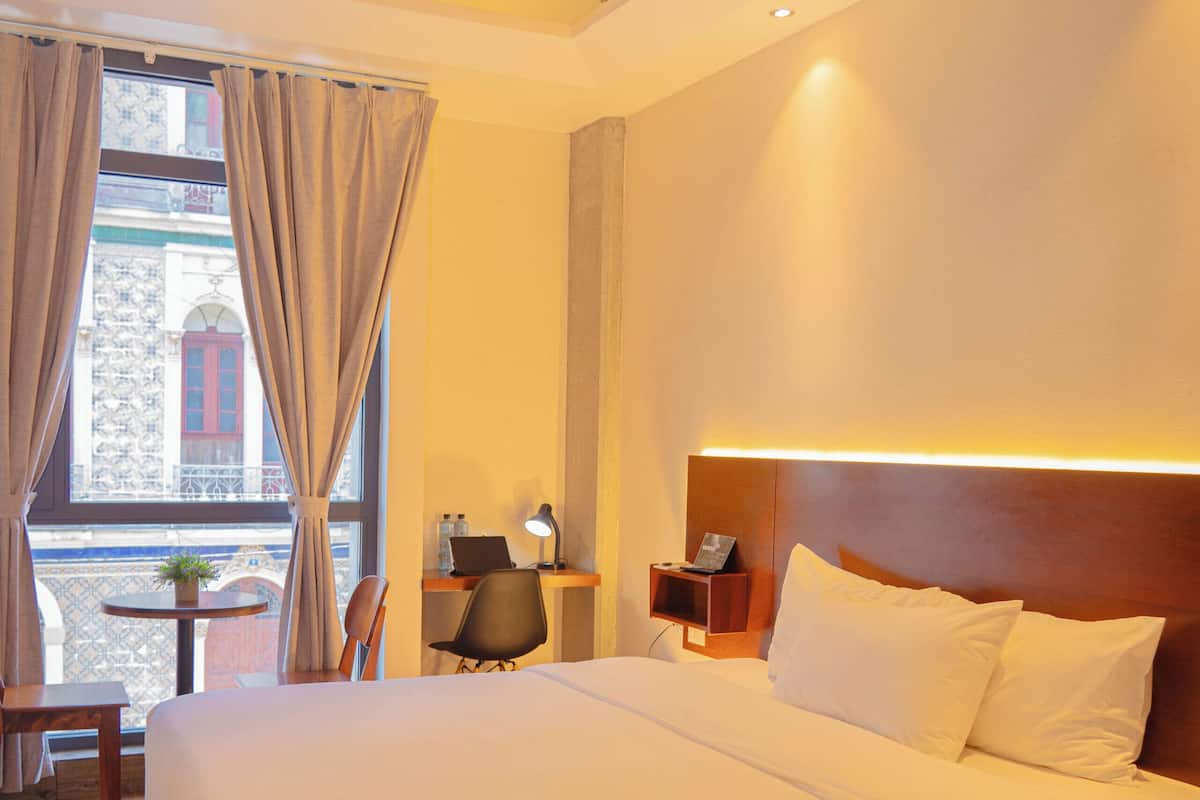 Une chambre d’hôtel avec un grand lit, un bureau avec une chaise, une petite table avec une plante et une vue sur un immeuble à travers une fenêtre avec des rideaux.