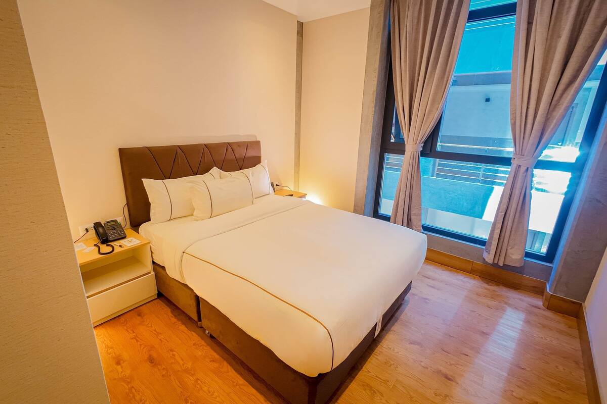 Une chambre d’hôtel avec un grand lit, une table de chevet avec un téléphone et une fenêtre avec des rideaux.