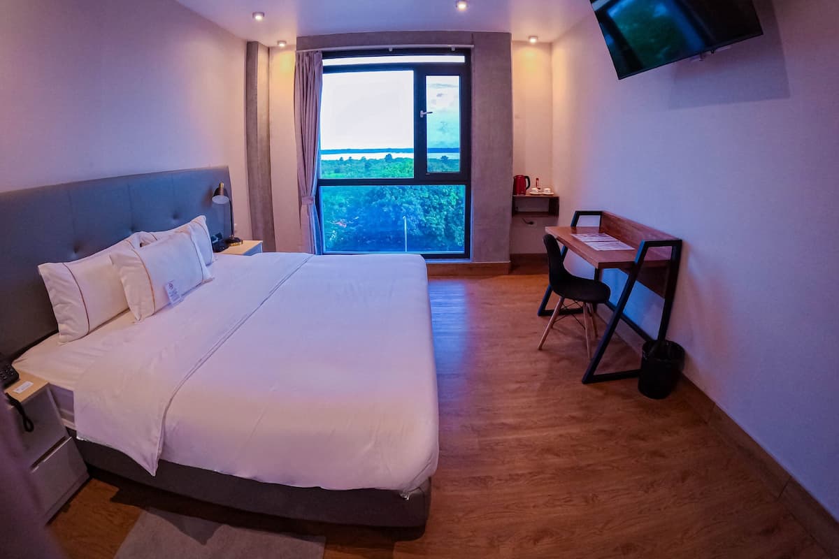 Une chambre d’hôtel avec un grand lit, un bureau avec deux chaises, un téléviseur fixé au mur et une fenêtre donnant sur un paysage verdoyant.