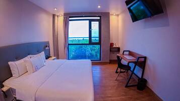 Quarto de hotel com uma cama grande, uma escrivaninha com duas cadeiras, uma televisão na parede e uma janela com vista para áreas verdes.