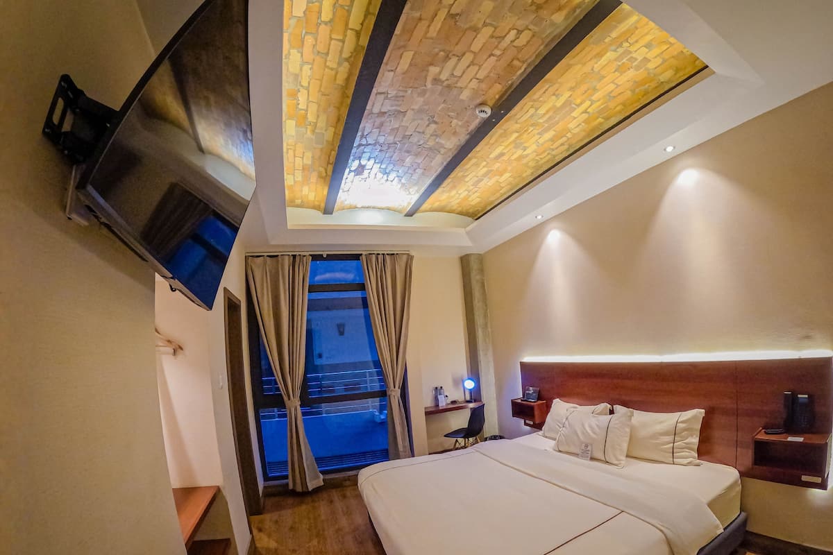 Une chambre d’hôtel avec un grand lit, un téléviseur à écran plat fixé au mur, une table de chevet avec une lampe et une vue sur l’extérieur par une fenêtre munie de rideaux.