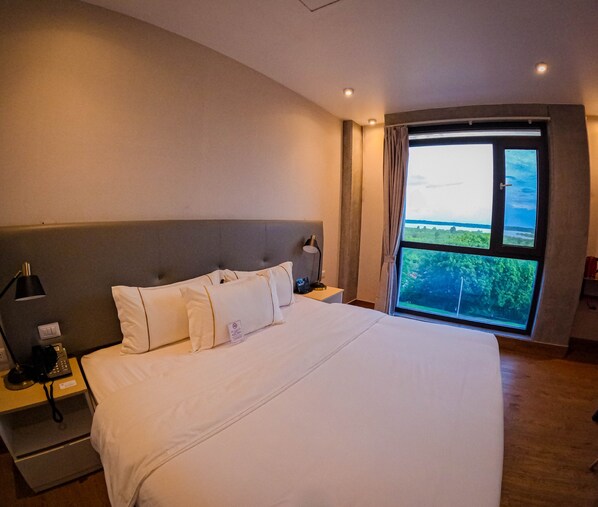 A large bed with white linens, a grey upholstered headboard, bedside lamps, a nightstand with a phone, and a window with a view of greenery.