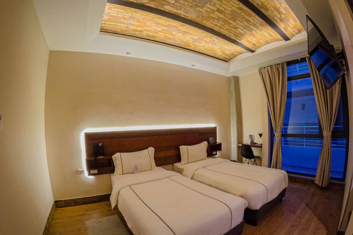 Une chambre d’hôtel moderne équipée de deux lits, d’un bureau, d’une chaise et d’une grande fenêtre avec des rideaux.