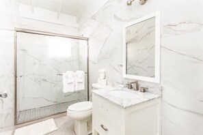 Une salle de bain moderne avec une douche à l’italienne en verre, des toilettes blanches et un meuble-lavabo en marbre agrémenté d’un grand miroir.