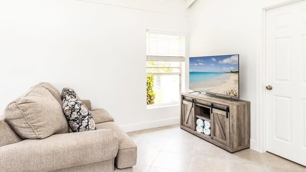 Een woonkamer met een beige bank, een televisie op een houten dressoir en uitzicht op een strand door een raam.
