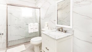Une salle de bain moderne avec une douche à l’italienne en verre, des toilettes blanches et un meuble-lavabo en marbre agrémenté d’un grand miroir.
