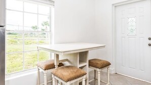 Een kamer met een witte tafel en drie bijpassende krukken, een groot raam met uitzicht op groen en een witte deur met een decoratief glaspaneel.