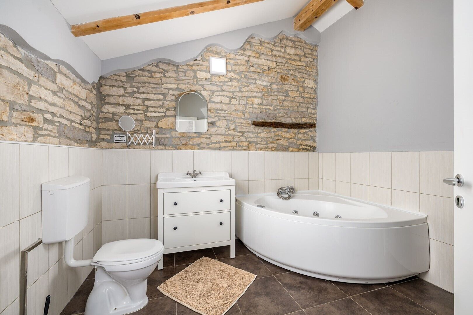A bathroom with a stone wall, a white toilet, a vanity with drawers, a bathtub, and a wooden ceiling beam.