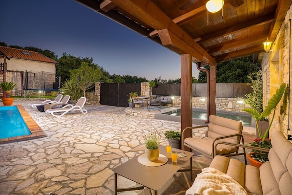 A stone patio with a pool, lounge chairs, and a covered seating area with a table and chairs.