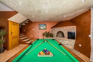 Una mesa de billar con superficie de fieltro verde, una chimenea de ladrillo y una escalera que sube a un nivel superior.