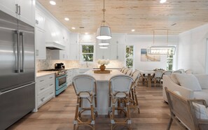 Una cucina moderna con mobili bianchi, un frigorifero in acciaio inossidabile, un forno blu e un'area pranzo con sedie in vimini e un tavolo bianco.