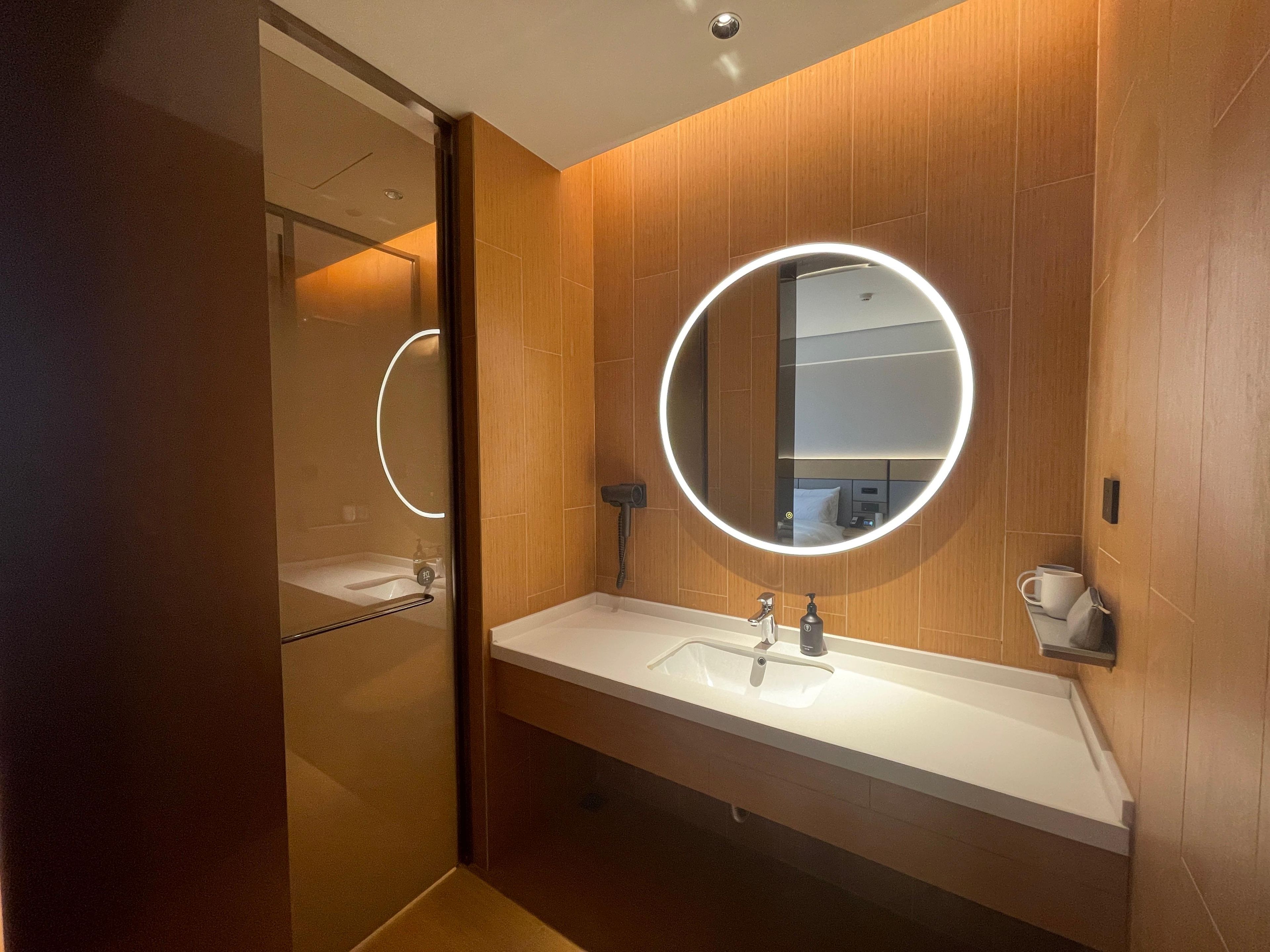 A modern bathroom with a wooden wall, a round mirror with a light, a white sink, and a shower area.