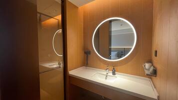 A modern bathroom with a wooden wall, a round mirror with a light, a white sink, and a shower area.