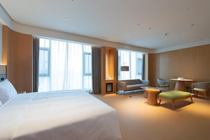 Una habitación de hotel moderna con una cama grande, una zona de estar con sofá y sillones, una mesita y ventanas amplias con cortinas.
