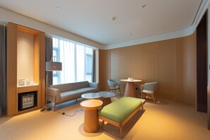 Quarto de hotel moderno com sofá, duas mesas redondas, uma mesinha de centro, televisão e minibar.