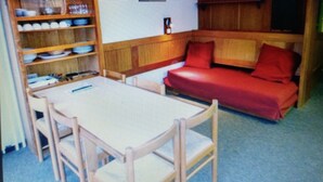 A room with a wooden table, chairs, a red sofa, and a bookshelf with various items. - SKI IN THE SUN: February 16 to 23, 2025, Serre Chevalier Chantemerle club Maeva (Saint-Chaffrey)