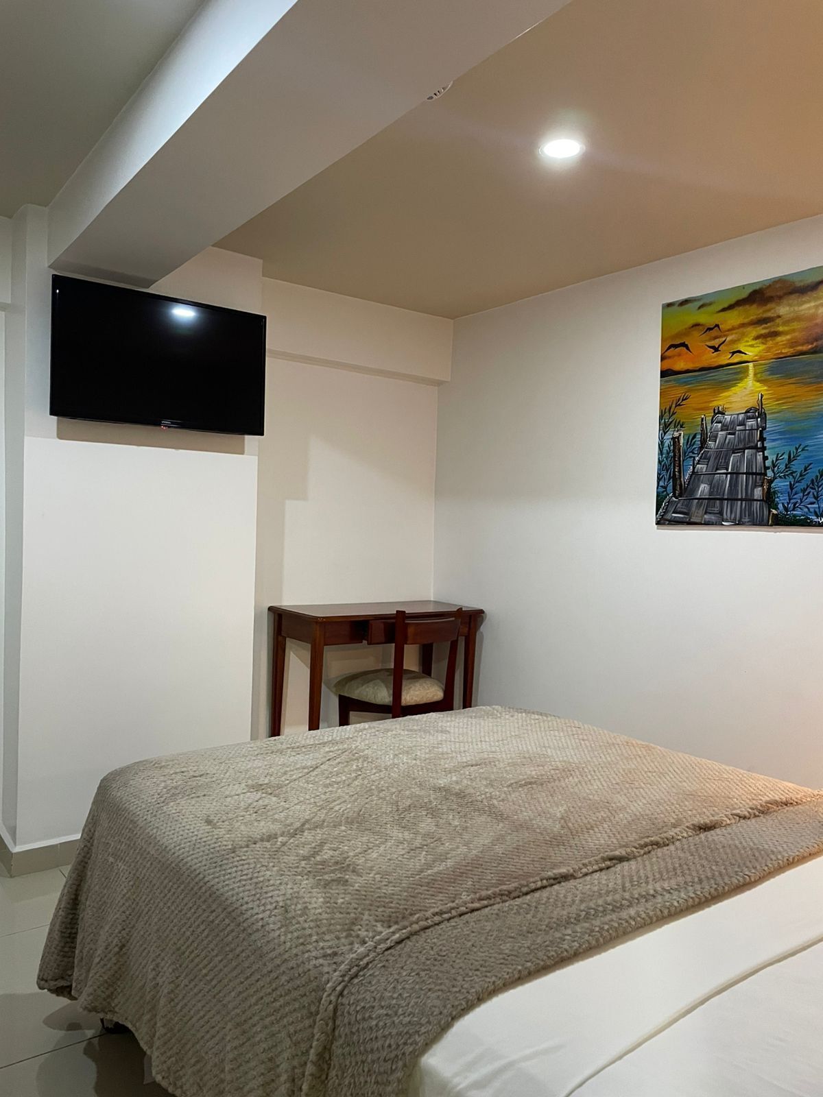 A bedroom with a bed, a desk, a chair, a television, and a painting on the wall.