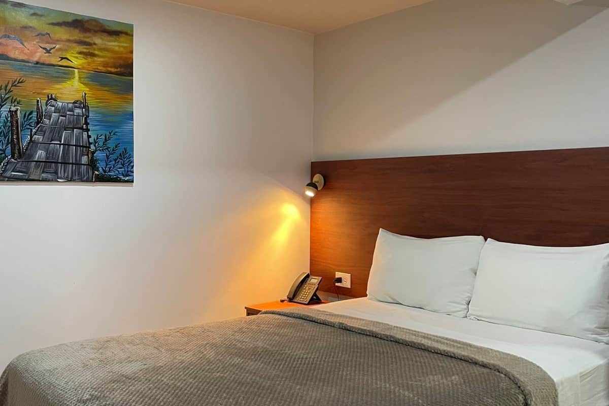 A hotel room with a bed, a nightstand with a phone, a painting of a sunset over a dock, and a wooden headboard.