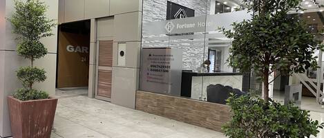 A hotel reception area with a modern design, including a reception desk, a glass wall, and a signboard with contact information.