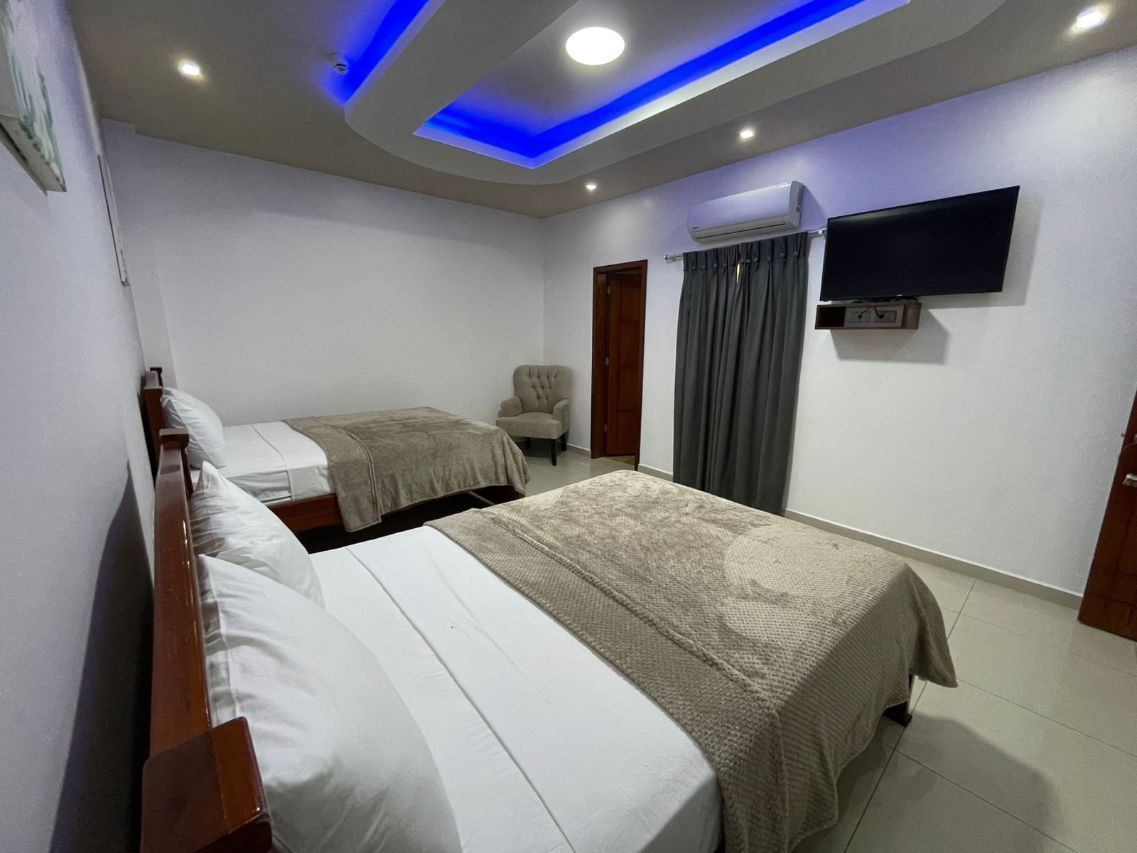 A hotel room with two beds, a flat-screen TV mounted on the wall, an air conditioning unit, and a ceiling with blue lighting.