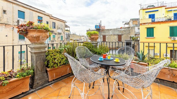 Une terrasse sur le toit, avec une table ronde et des chaises en treillis métallique, entourée de jardinières en terre cuite où sont plantées différentes espèces végétales.