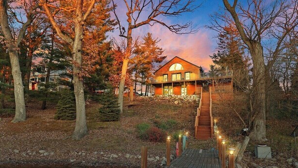 A wooden house with a stone staircase leading to a deck, surrounded by trees and a small stream. - Woodhaven on Pleasant Lake |Pleasant Lake |Lakefront |Wraparound Deck |Views |Dock |Kayaks & Boards (East Troy)