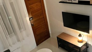 A modern living room with a flat-screen TV, a wooden door, a white round coffee table, and a black sofa with patterned cushions. - 2BR Home (corner lot). With Lanai, 100 Mbps Internet and Netflix. (Balanga)